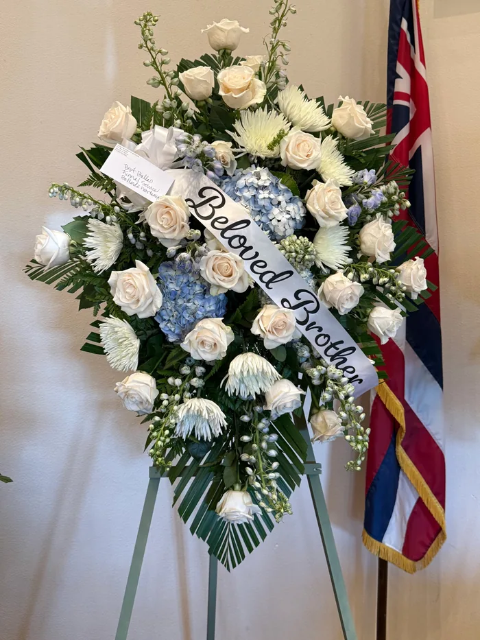 A blue and white standing funeral spray with a hand lettered ribbon sash.