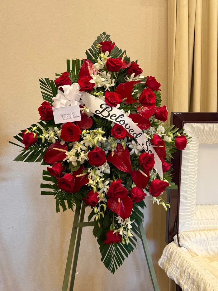 A standing funeral spray of deep red roses with palm frond and fern greenery.