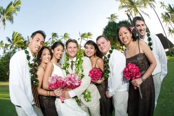 A wedding party photographed outdoors, the men wearing lei and the women holding pink rose bouquets.