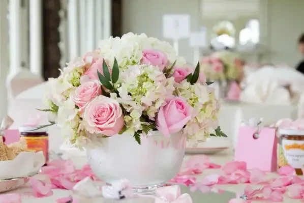 A footed bowl of pink roses and white hydrangea on a table scattered with petals.